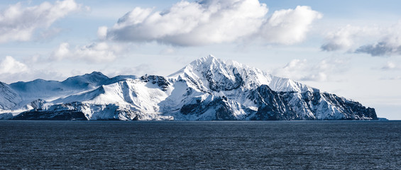 Fototapeta na wymiar Snow peaks, glaciers and rocks of Aleutian islands in sunny winter day as viewed from ship passing in calm sea