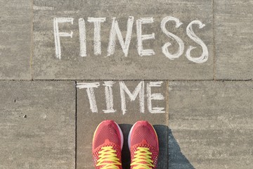 Text fitness time written on gray pavement with woman legs in sneakers, view from above