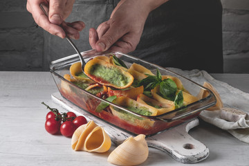 Cooking by chef hands traditional pasta recipe with greens, herbs, tomatoes and cheese, baked in glass tray on white wooden background.