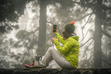 Fototapeta na wymiar Mountain trekking lover woman makes a break drinking hot coffee and making a video call with remote friends. Sporty girl sitting on a wall in a misty forest shooting selfies. Winter autumn landscape