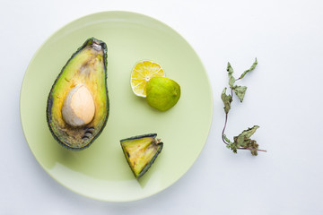 Ugly food on white background. Ugly rotten avocado on green plate. Bad lime and dry mint on a plate. Rotten tropical fruits. Concept of rotten fruit. Top view, copy space