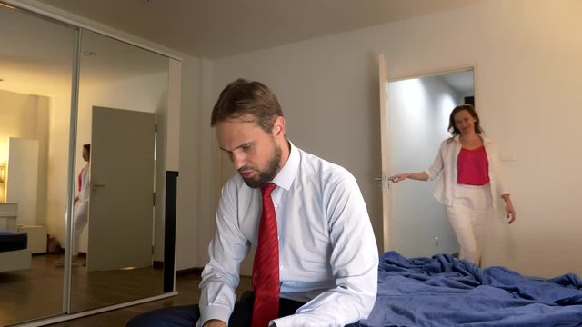 tired man sitting on the bed after work day. An attractive charming seductive woman enters the bedroom and whispers pleasant words to her sleepy tired man.
