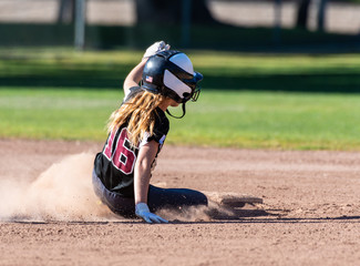 Female teenage softball player in black uniform sliding safely into second base.