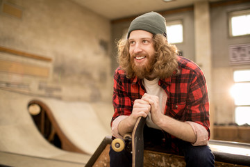 Waist up portrait of long haired skater sitting on ramp in extreme park and smiling, copy space