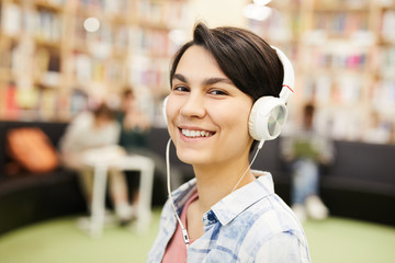 Smiling beautiful hipster girl listening to audiobook in headphones and looking at camera while spending time in library