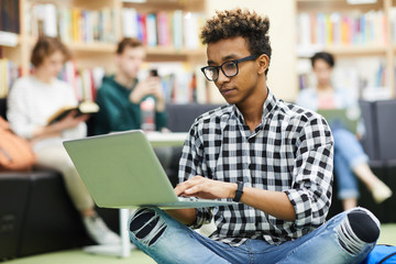 Serious thoughtful young black student boy in glasses sitting with crossed legs in library and preparing research for university project