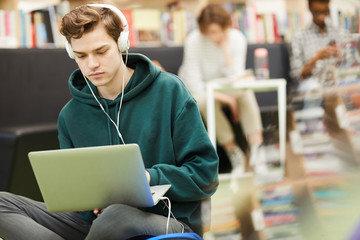 Serious hipster student boy listening to music in headphones and using laptop while searching for...