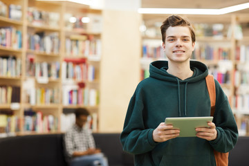 Portrait of smiling modern student guy in dark green hoodie wearing satchel on shoulder standing in library and using gadget