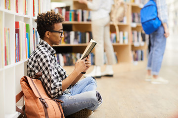 Serious clever boy with Afro hairstyle sitting with crossed legs on floor in library and reading...
