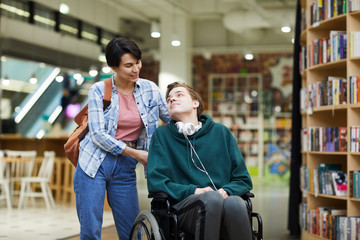 Positive attractive young woman with satchel pushing wheelchair and talking to disabled student in...