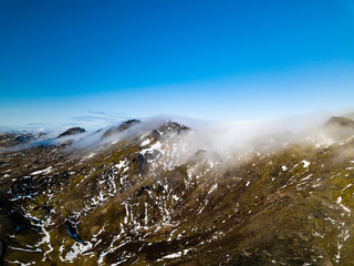 Drone photo of the top of a mountain chain covered in clouds and illuminated by the morning sun 
