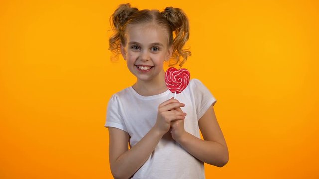 Pretty candy girl licking delicious heart-shaped lollipop and smiling, happiness