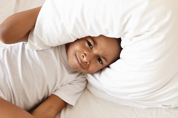 Bedtime, comfort and bedding concept. Top view of frustrated upset little black boy wearing white...