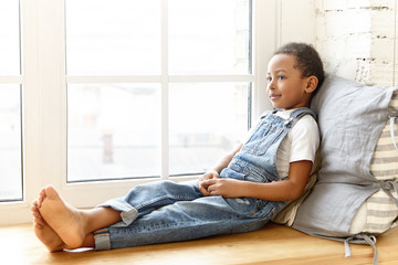 Indoor portrait of charming cute eight year old dark skinned African schoolboy wearing jeans jumpsuit spending nice time at home after school, sitting barefooted on windowsill and smiling happily