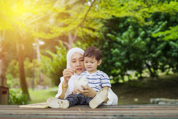 Muslim Asian young mother with her baby son, learning and putting coins into piggy bank at park. Education, financial and future planning concept.