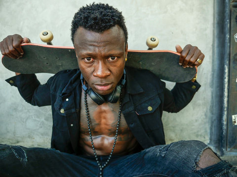 cool isolated portrait of young attractive and confident hipster black afro American man leaning on street wall holding skate board as badass guy in urban style