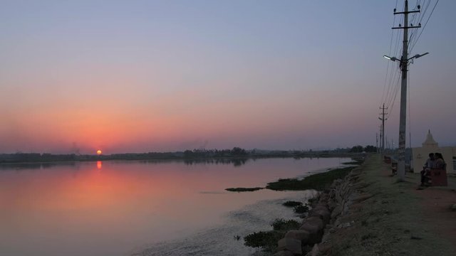 People seated at a lake side as they watch the sunset in a romantic setting with the reflection in the water in Karnataka, India 