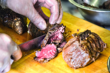 Chef Hands in Disposable Food Preparation Plastic Gloves Carving Cooked Roast Beef Sirloin with a Big Knife on a Wooden Cutting Board. Catering Services, Cooking Master Class, Workshop.
