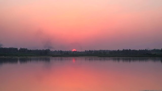 Time lapse of sun setting in a brilliant colorful red and orange sky with reflection in the water across the lake and forest somewhere on the highway in Karnataka, India 