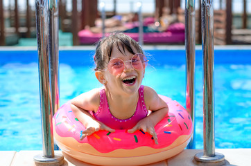 Cute laughing little girl with inflatable donut ring in swimming pool on hot sunny day. Healthy and happy childhood concept.