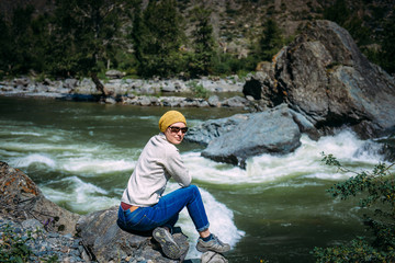 A young woman in sunglasses near the mountain river. Green river between the rocks and a girl sitting on the stone. A girl looking at camera, close-up. The river Chulyshman, Altai, Russia.