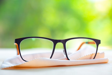 Black eyeglasses with ivory colour Microfibre cleaning cloths on wooden table, In bokeh green...