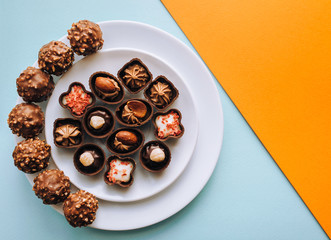 Beautiful chocolates of different shapes and fillings lie in a white plate against the background of colored paper. Orange and blue color. Copy space, diagonal.