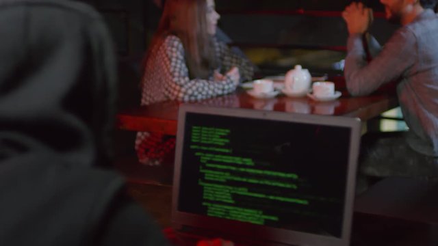 Rare view of young man sitting in bar at laptop and developing new software while people on background having meal