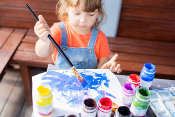 Adorable little kid girl holding paint brushes, painting picrure at the paper
