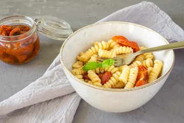 Fusilli pasta with dried tomatoes and basil in a bowl on a gray concrete background with copy space