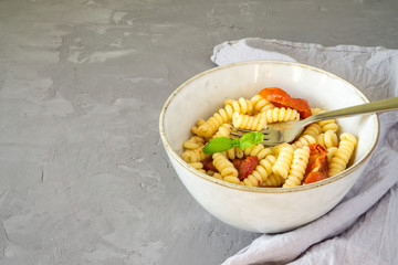 Fusilli pasta with dried tomatoes and basil in a bowl on a gray concrete background with copy space