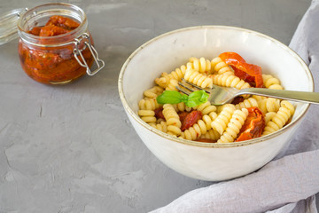 Fusilli pasta with dried tomatoes and basil in a bowl on a gray concrete background with copy space