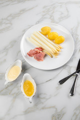 Weißer Spargel mit Sauce Hollandaise, zerlassener Butter, geräucherter Schinken und Kartoffeln auf Teller und Marmor Hintergrund