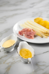 Weißer Spargel mit Sauce Hollandaise, zerlassener Butter, geräucherter Schinken und Kartoffeln auf Teller und Marmor Hintergrund