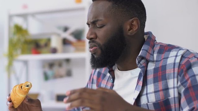 African american guy biting croissant and feeling toothpain, gum sensitivity