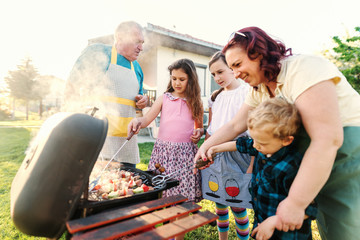 Mother teaching her little son how to grill meat on the grill. next to her girls and grandfather....