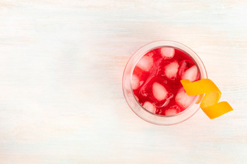 A vibrant red cocktail with campari and an orange curl garnish, shot from the top on a white wooden background with ice cubes and copy space