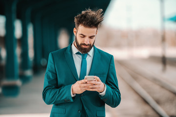 handsome Caucasian bearded businessman in turquoise suit using smart phone while waiting for train...