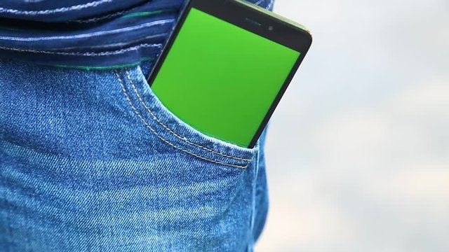 Closeup view of modern smartphone with empty blank green screen seen from out pocket of jeans of young kid standing outdoors. Real time full hd video footage.