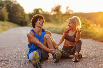 Fit cute couple sitting on the country road and resting from running. Man tying shoelace. Summer...