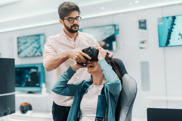 Cheerful multicultural couple trying out virtual reality technology in tech store. Man helping woman to put vr goggles and woman sitting in chair.