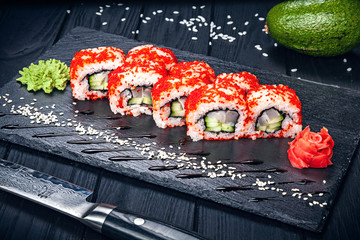 Close up view California roll sushi with tobiko caviar, sea bass, cream cheese on dark stone served with ginger and wasabi and black wooden table. Japanese cuisine. Copy space. Served sushi for menu