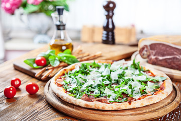 Selective focus on fresh pizza with prosciutto on a wooden table on light background. Pizza with ingridiens: cherry tomato, basil, olive oil. Horizontal image with copy space for text. Food for menu