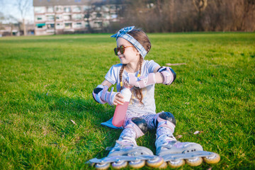 Cute girl without milk tooth and two braid sitting on grass in roller skates and sunglasses and open bottle with water on beautiful sunny day