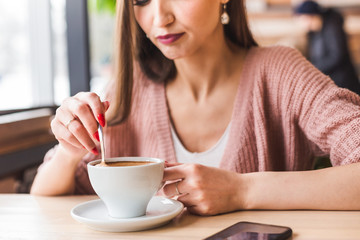 Attractive young woman sits at a table in a cafe with a cup of coffee and mixes it with a spoon