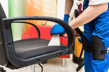 Young man in workwear and rubber gloves cleans the office chair with professional equipment.