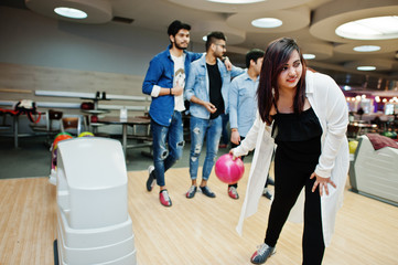 South asian woman standing at bowling alley with ball on hands. Girl is preparing for a throw. Friends support her loudly.