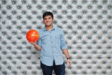 Stylish asian man in jeans shirt standing with bowling ball at hand against silver wall background.