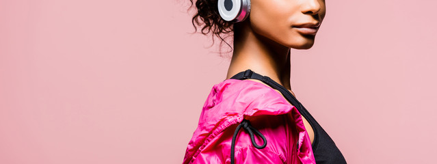 panoramic shot of african american sportswoman in windbreaker and headphones isolated on pink with...