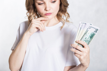 Pensive young woman looks at bunch of money, thinks where to spend it, over white background.